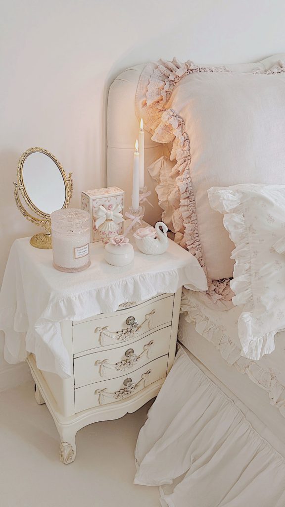 Romantic shabby chic nightstand styling featuring LoveShackFancy perfume, a La Jolie Muse scented candle, a vintage gold mirror, and a swan vase by La-Vania.