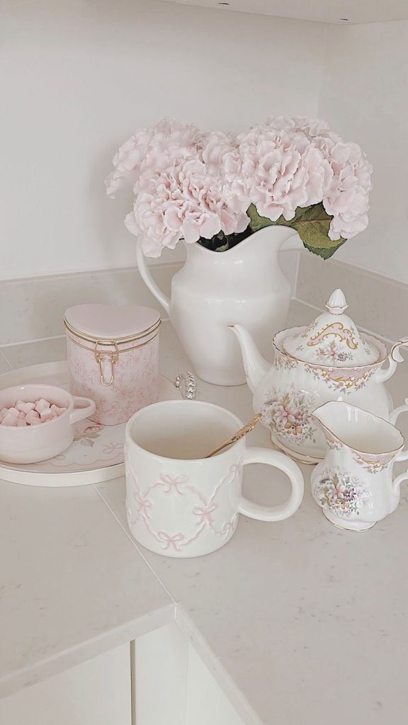 Shabby chic kitchen styling with a Royal Albert floral teapot, milk jar, pink bow mug, and heart-shaped hot chocolate tin on a marble countertop by La-Vania
